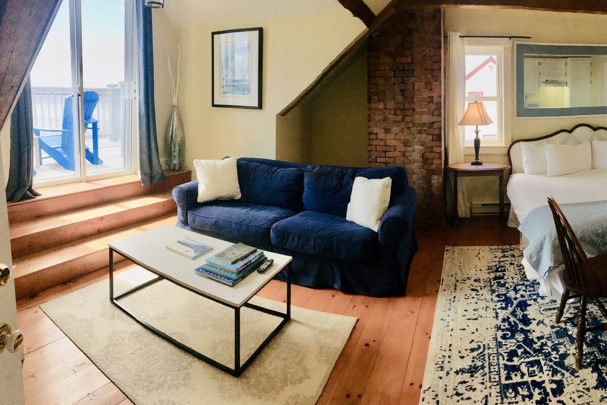 A cozy bedroom with a bed, a blue sofa, a small table, and a chair. There is a window with curtains, a mirror, and a lamp.