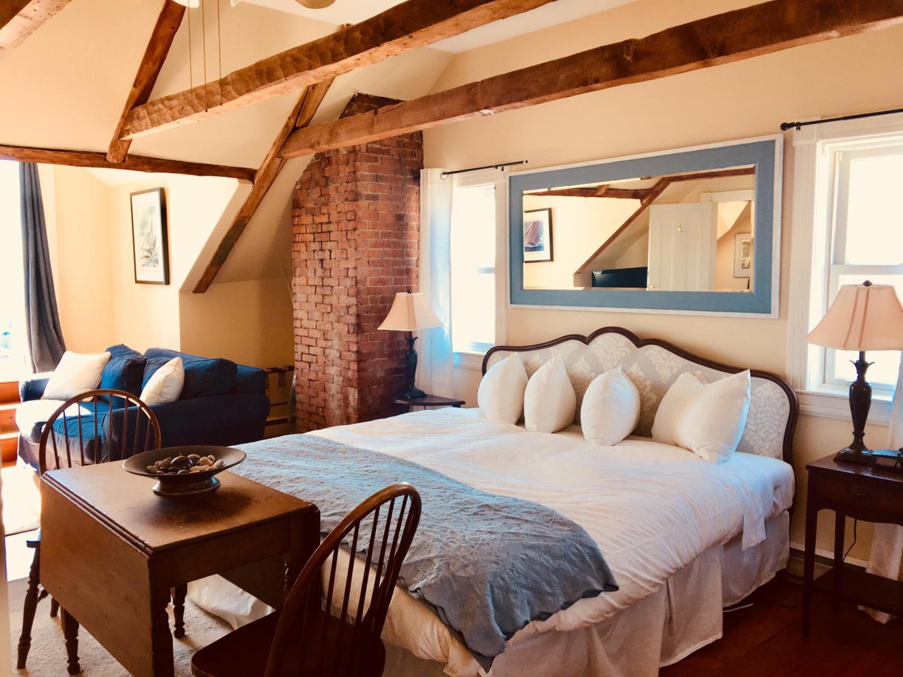 A bedroom with a large bed, a wooden table, a chair, a brick wall, and a window with a view.