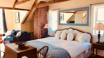 A bedroom with a large bed, a wooden table, a chair, a brick wall, and a window with a view.