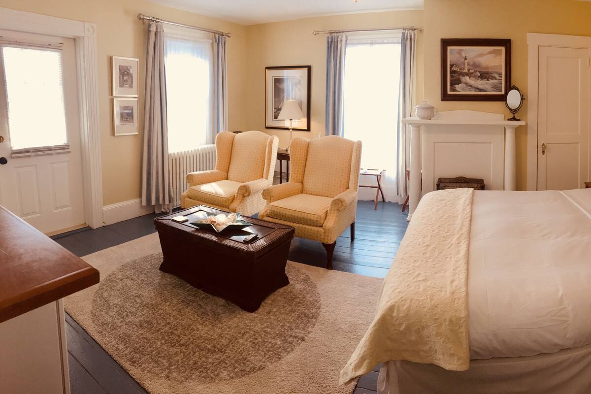 A hotel room with a bed, two armchairs, a coffee table, a fireplace, and framed artwork on the walls.