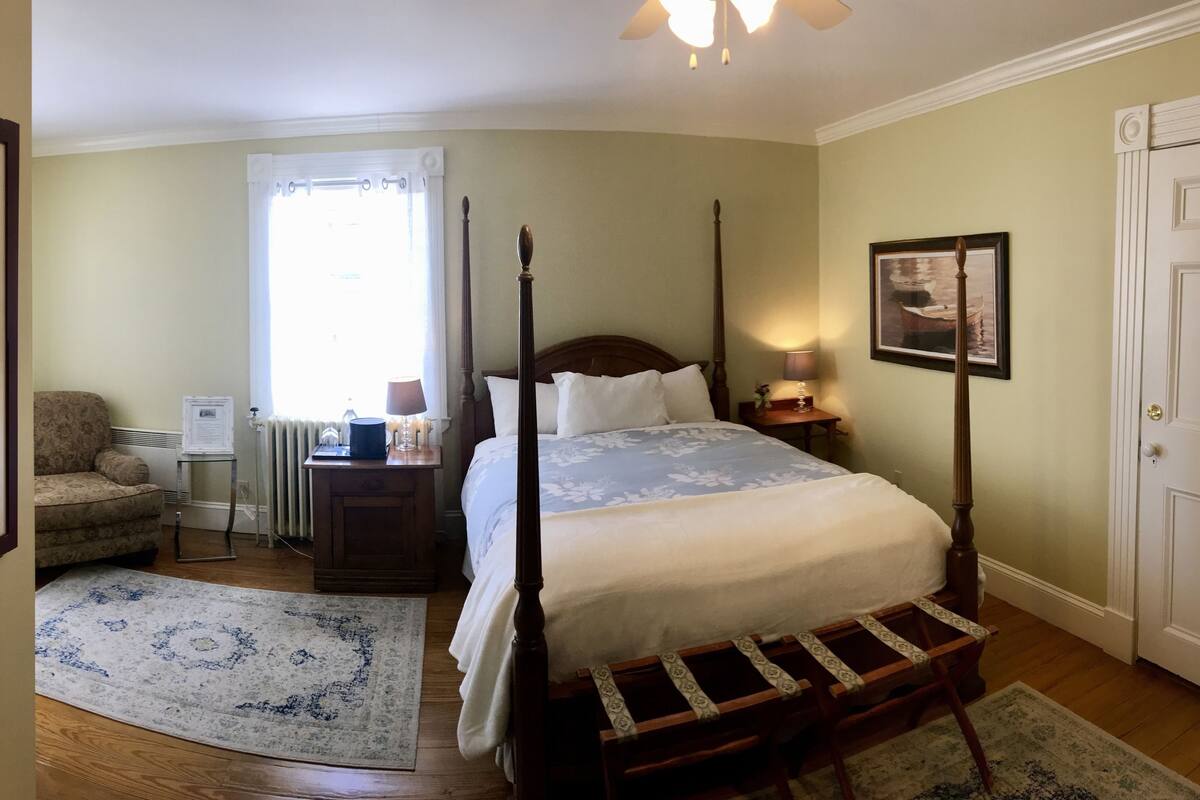 A bedroom with a four-poster bed, a nightstand, a chair, a radiator, a window with curtains, and a door.