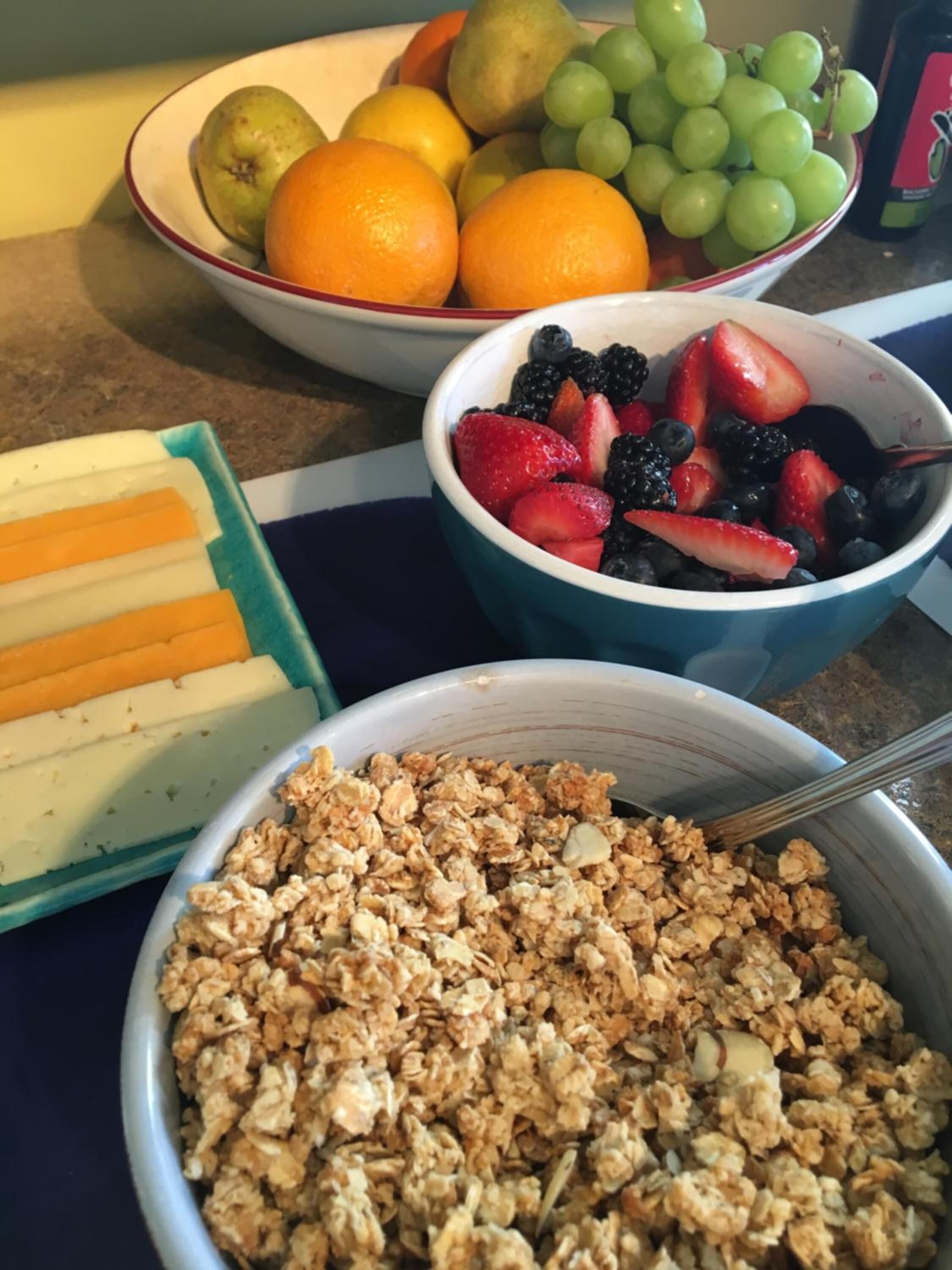 A breakfast table with bowls of granola, fresh fruits, and sliced cheese.