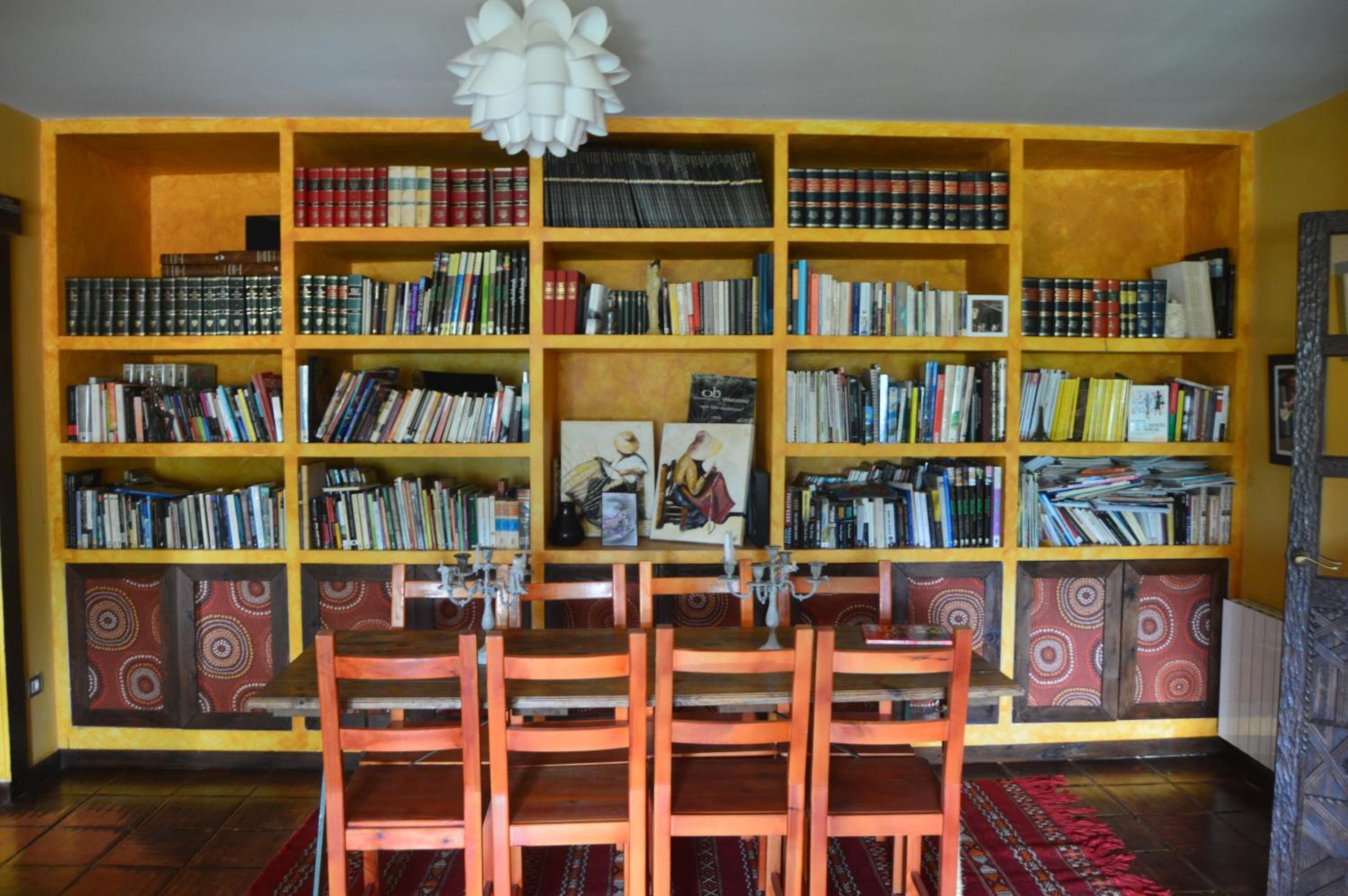 A room with a large bookshelf filled with books, a wooden dining table with chairs, and patterned wall panels.