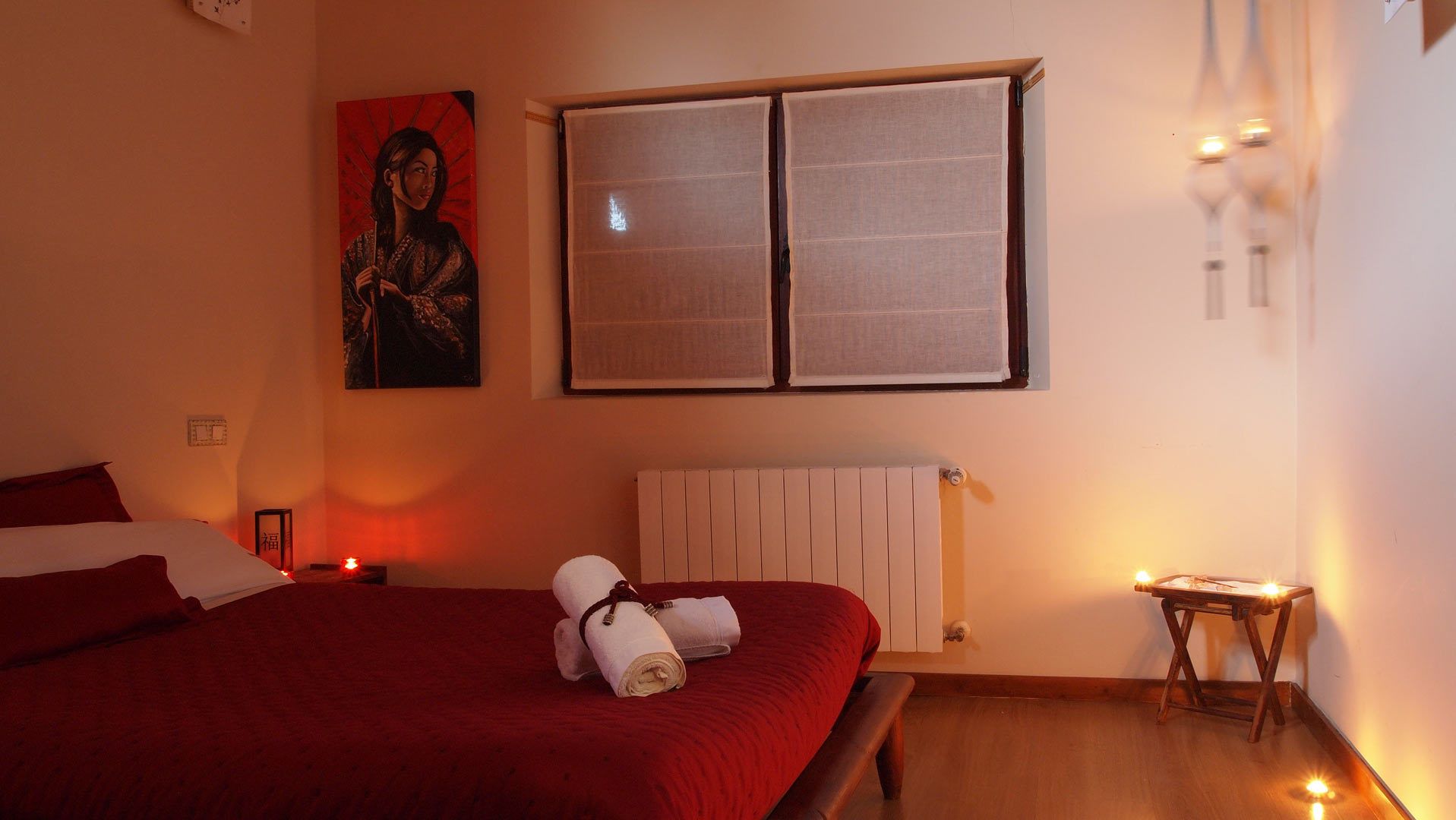 A bedroom with a bed, a window with blinds, a radiator, and a small table with a lamp.