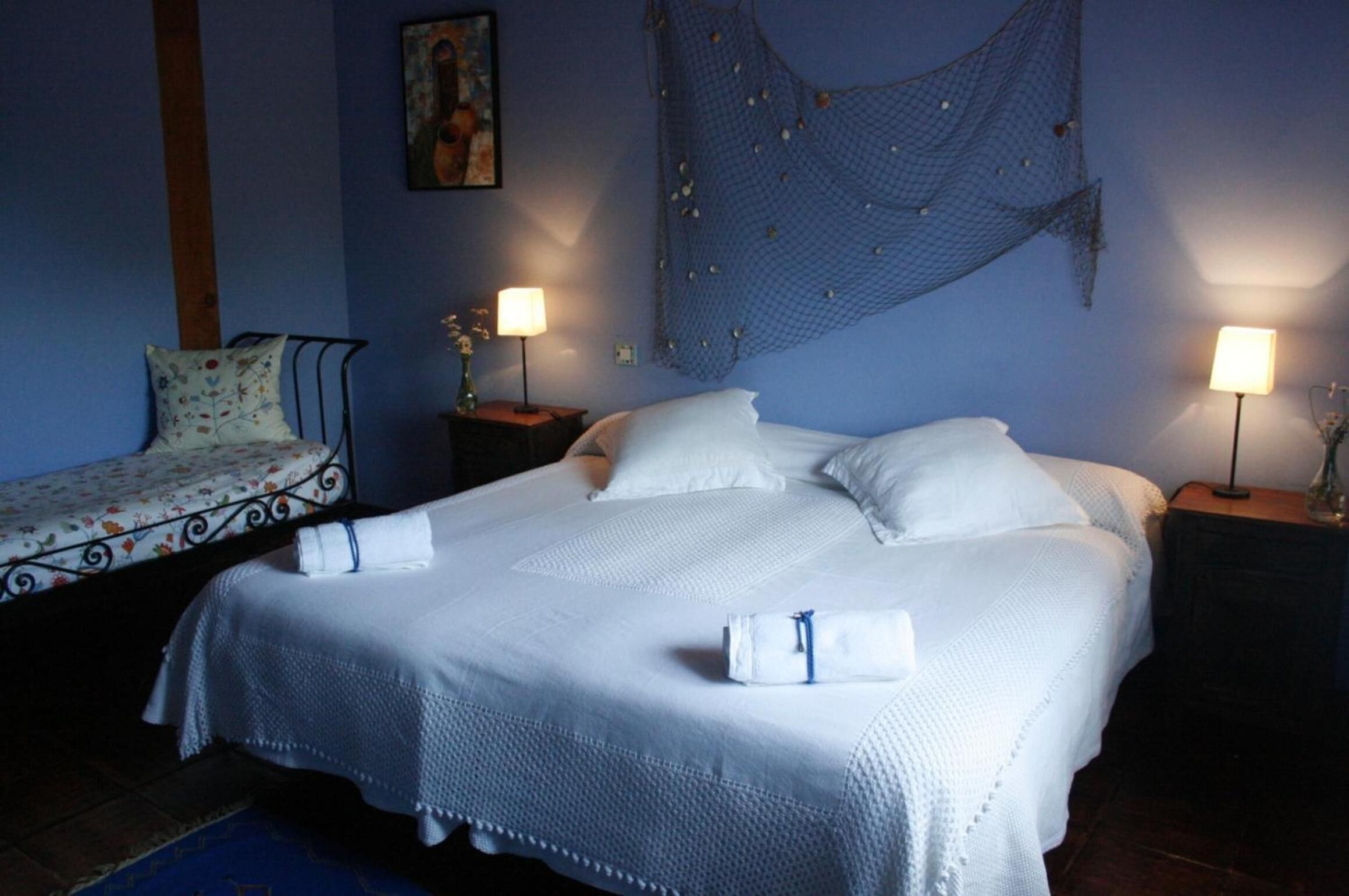 Una cama bien hecha con sábanas blancas, dos mesitas de noche con lámparas, una silla pequeña con cojín floral y un tapiz en la pared.