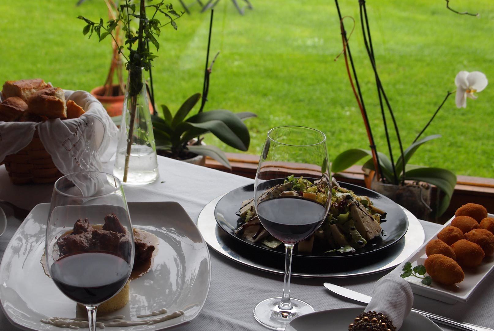 Una mesa dispuesta con un plato de comida, dos copas de vino tinto y una cesta de pan.