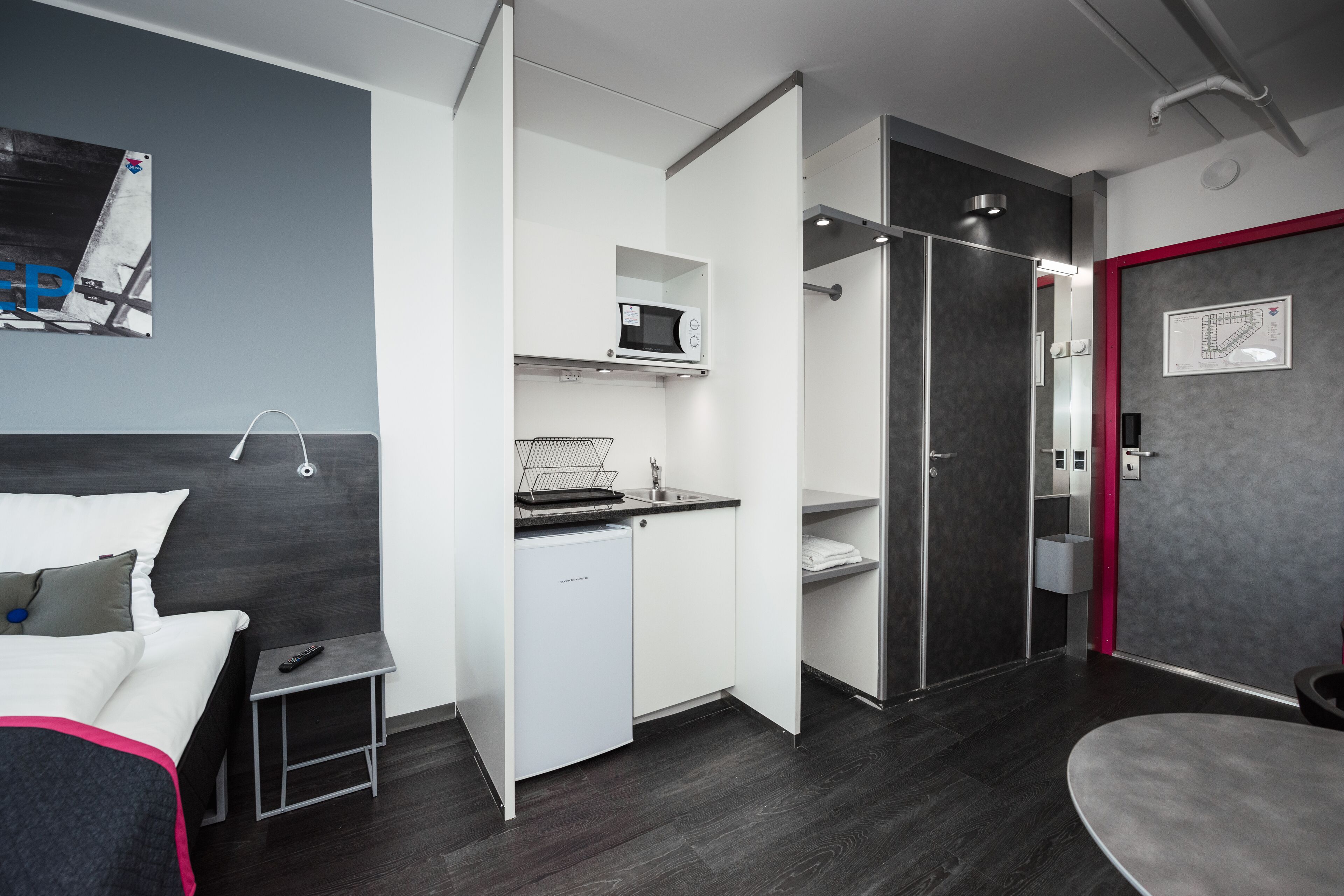 A modern hotel room with a bed, a small kitchenette, a bathroom, and a seating area.