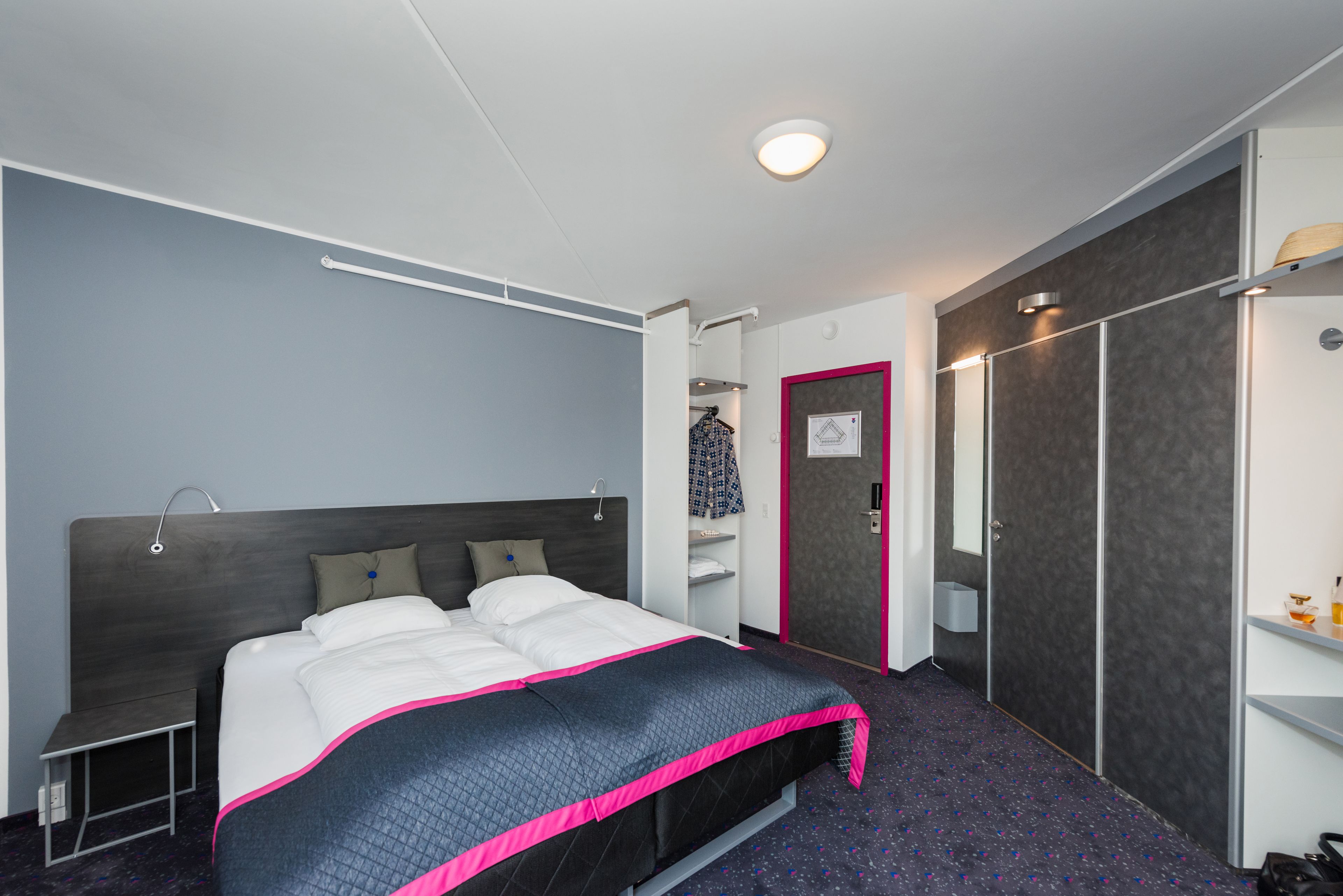 A modern hotel room with a bed, bedside table, wardrobe, and a small shelf with toiletries.