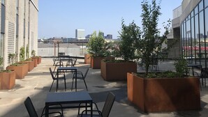 Un área de estar al aire libre con mesas y sillas, rodeada de macetas con follaje, y vistas del horizonte urbano.