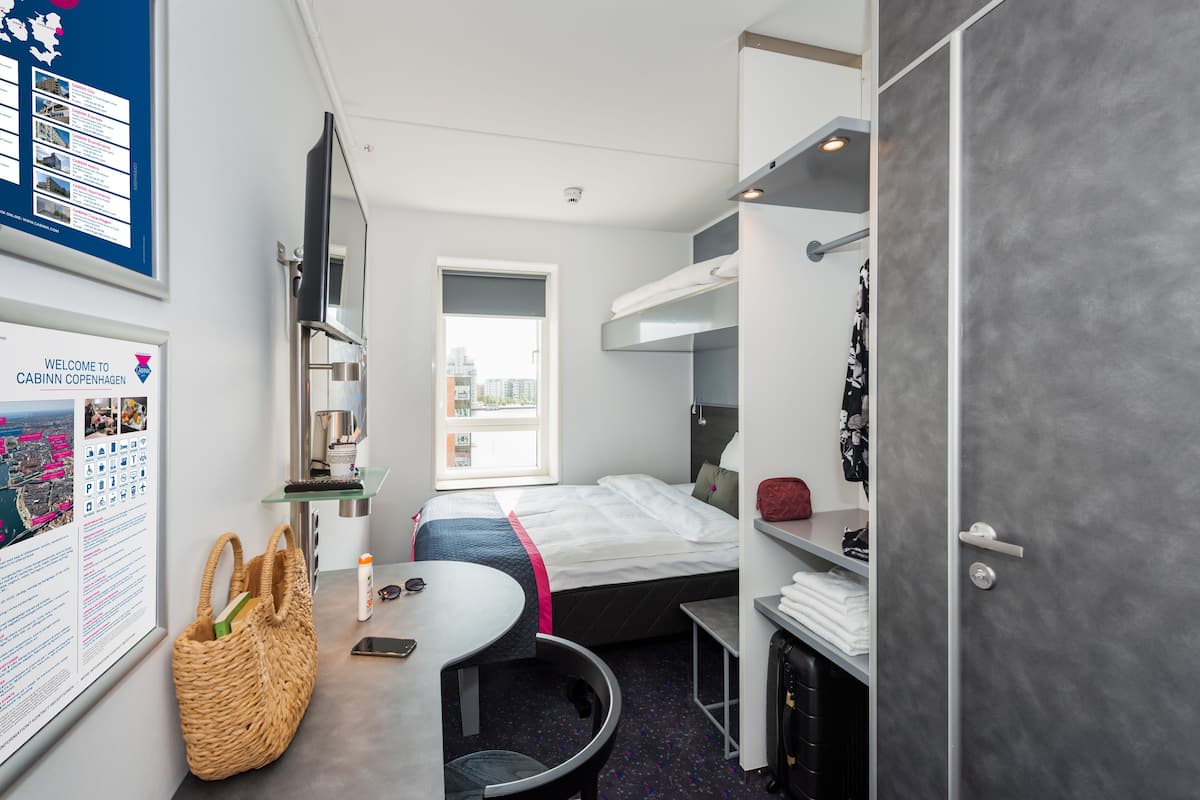 A modern hotel room with a bed, a desk, a chair, a small table, a TV, a window, and a wall-mounted shelf.