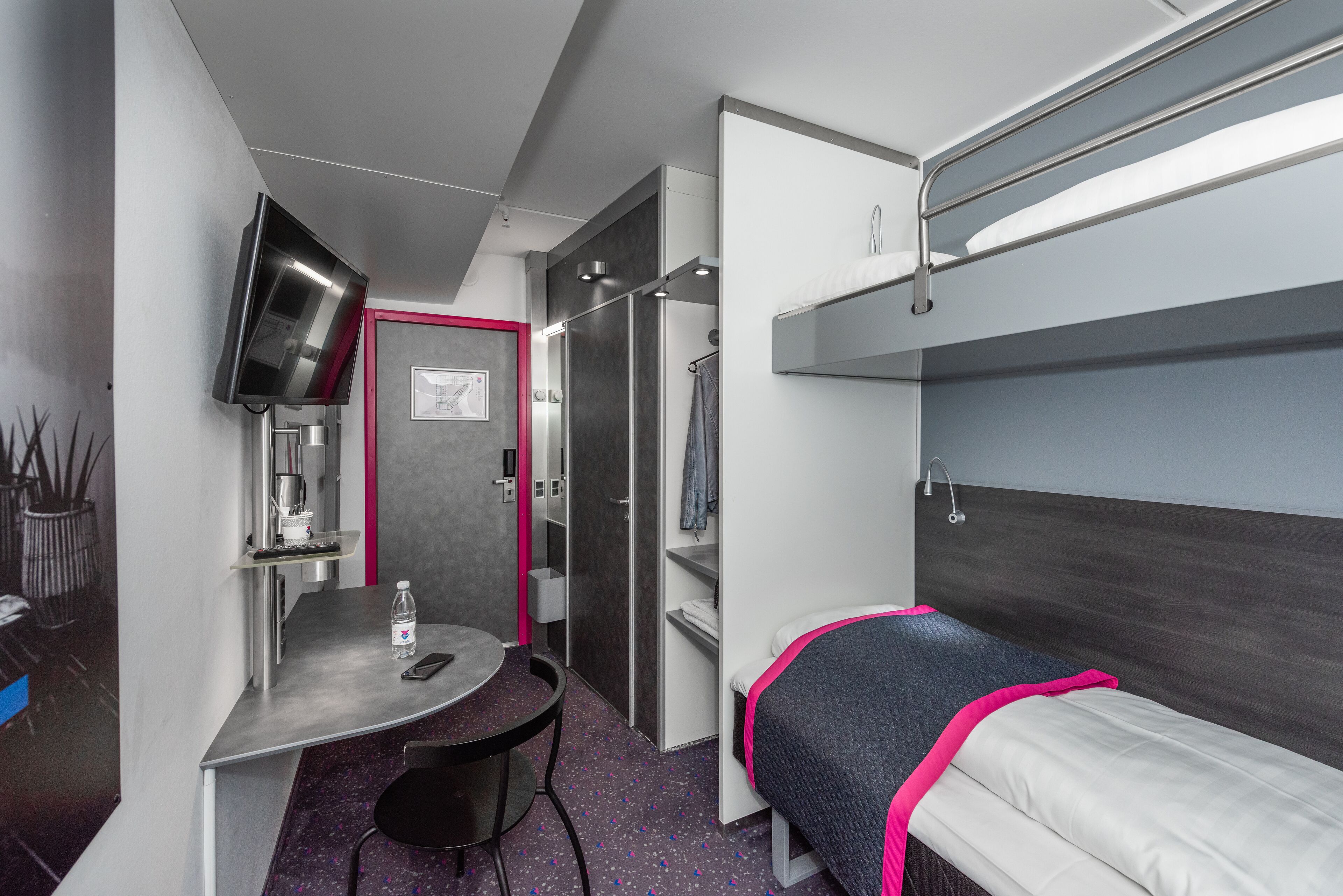 Habitación de hotel compacta con literas, un escritorio con silla, una mesita, televisor y zona de ducha.