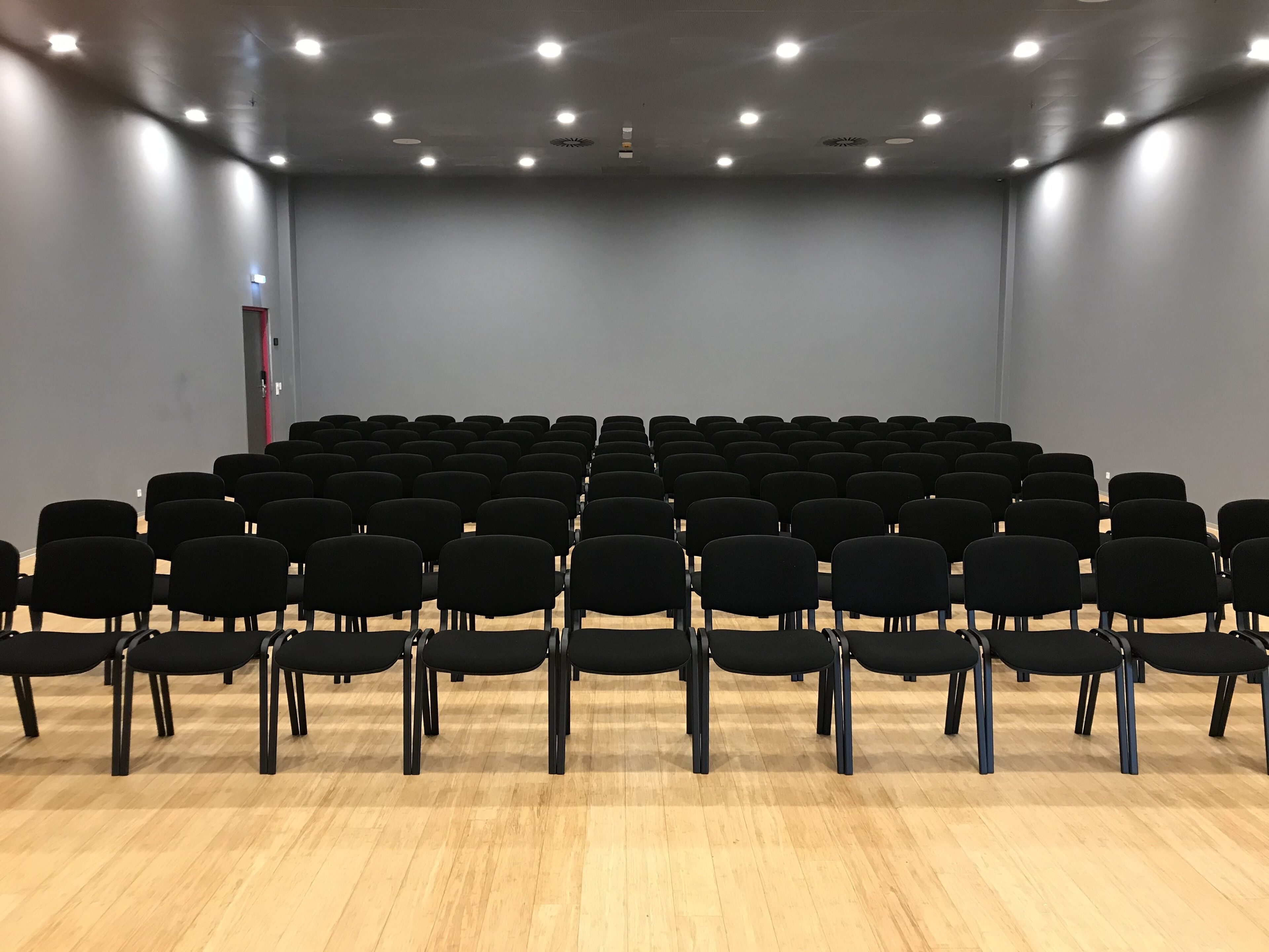 A conference room with rows of black chairs, a wooden floor, and a grey wall.