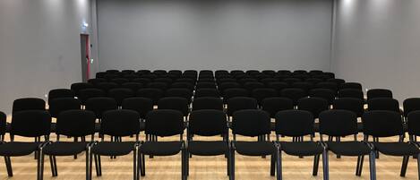 Une salle de conférence avec des rangées de chaises noires, un plancher en bois et un mur gris.