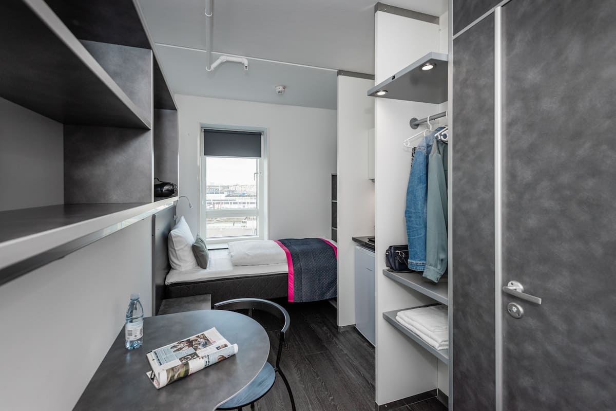 A compact room with a bed, a small table with a magazine, a water bottle, and a view of the city through a window.