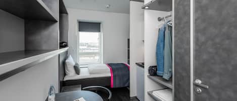 Une petite chambre comprenant un lit, une petite table avec un magazine, une bouteille d’eau et une vue sur la ville par la fenêtre.