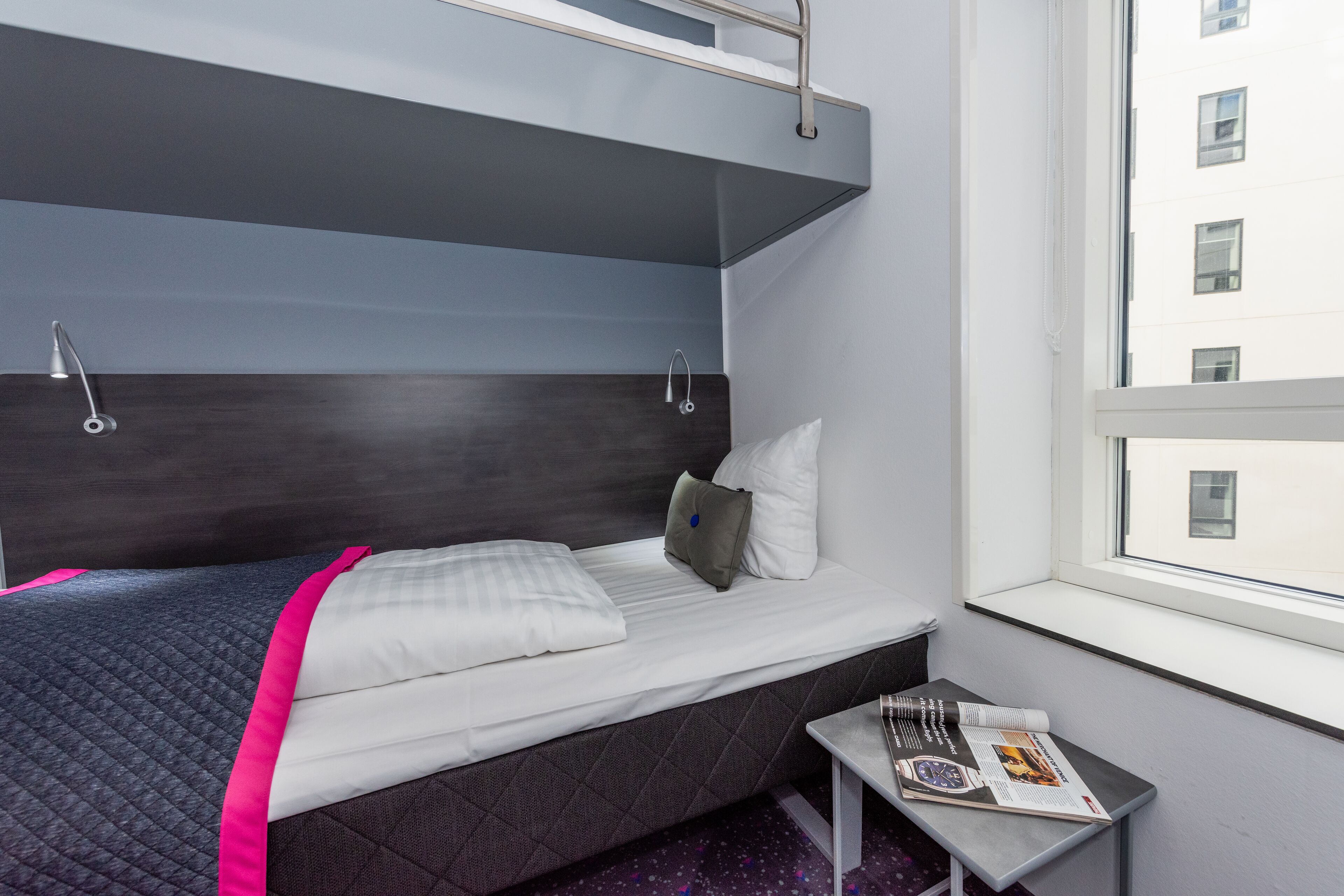 Een enkel bed met hoofdeinde, een klein tafeltje met een tijdschrift, en een raam met uitzicht op gebouwen.