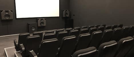A movie theater with rows of black seats, a large screen, and speakers on the walls.
