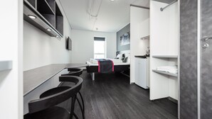 Una habitación de hotel moderna con kitchenette, una cama con una manta roja, un baño con toallero y un ventanal grande.
