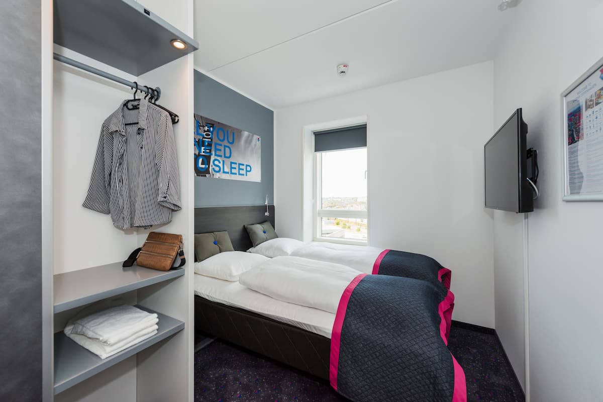 A hotel room with a bed, a wardrobe, a shelf with folded towels, a pillow, a bedside table, and a window.