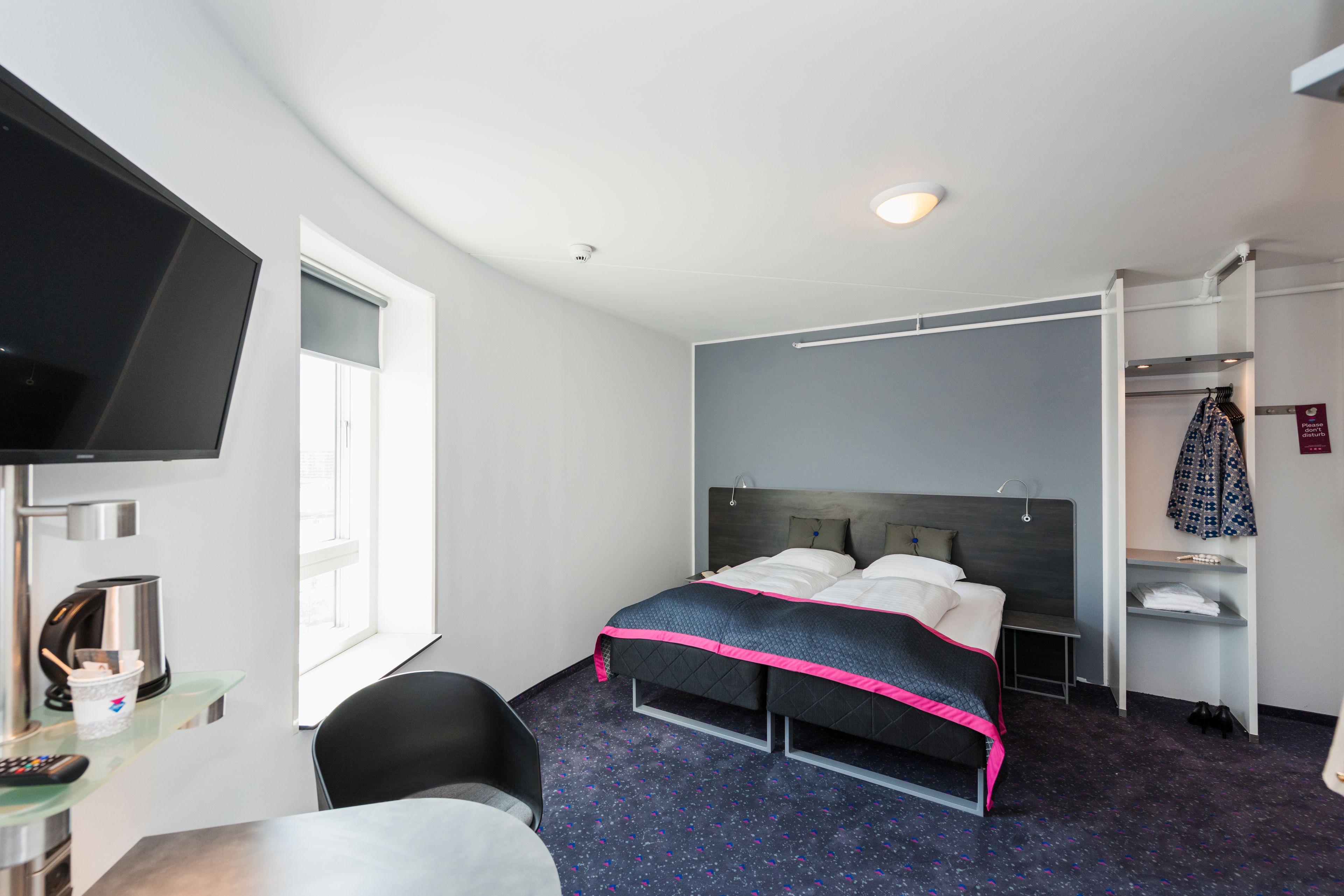 A hotel room with a bed, a TV, a desk, a chair, a shelf with clothes, and a window.