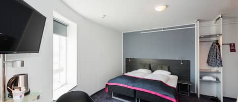 Habitación de hotel con cama, televisión, escritorio, silla, estante con ropa y ventana.
