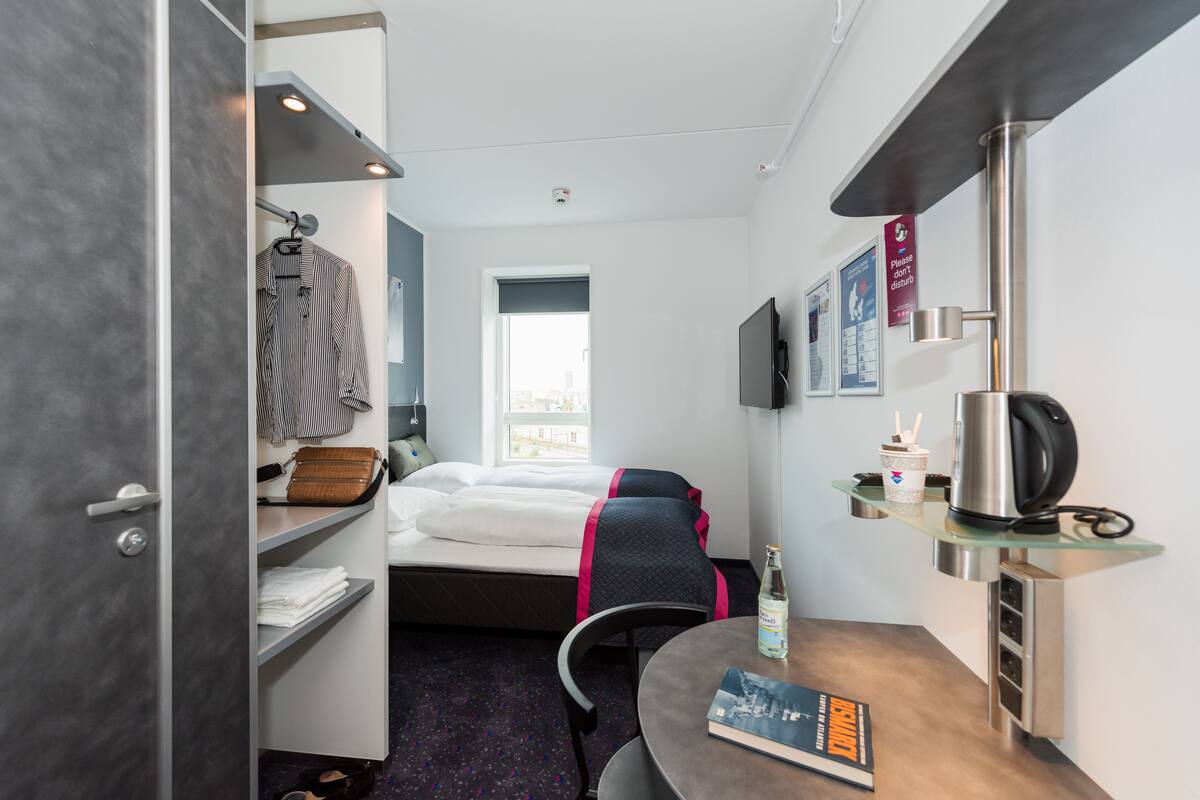 A compact hotel room with a bed, a small table with a book, a kettle, and a shelf with a bottle and a cup.
