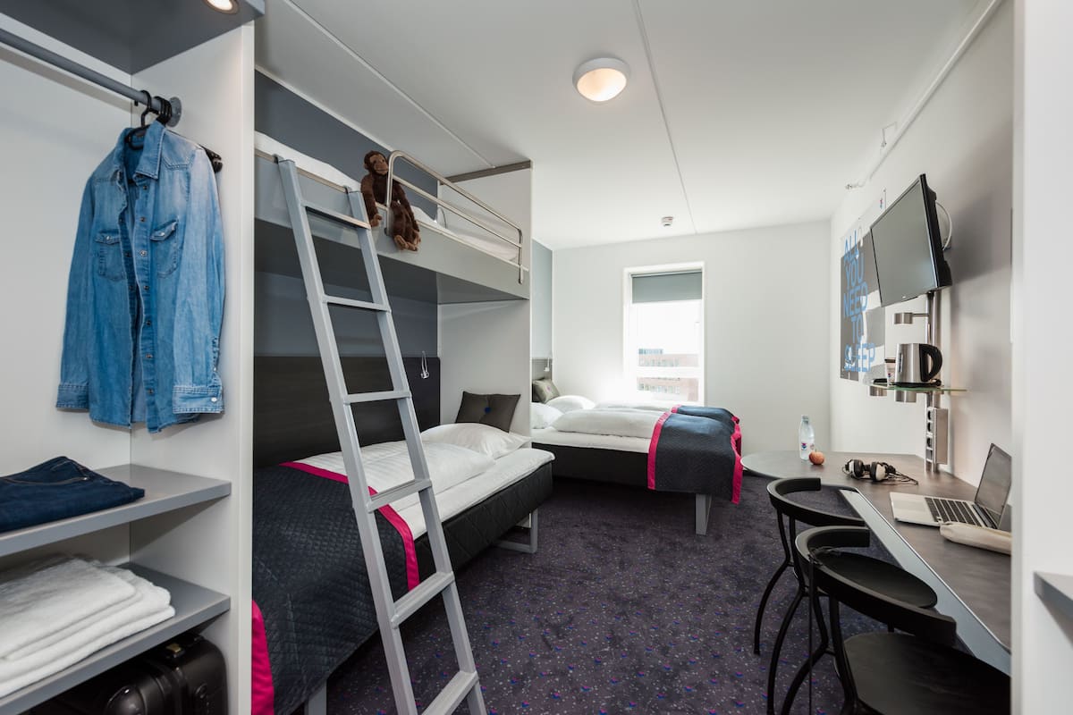 A dormitory room with bunk beds, a desk with a laptop, a chair, a ladder, a hanging jacket, and a window.