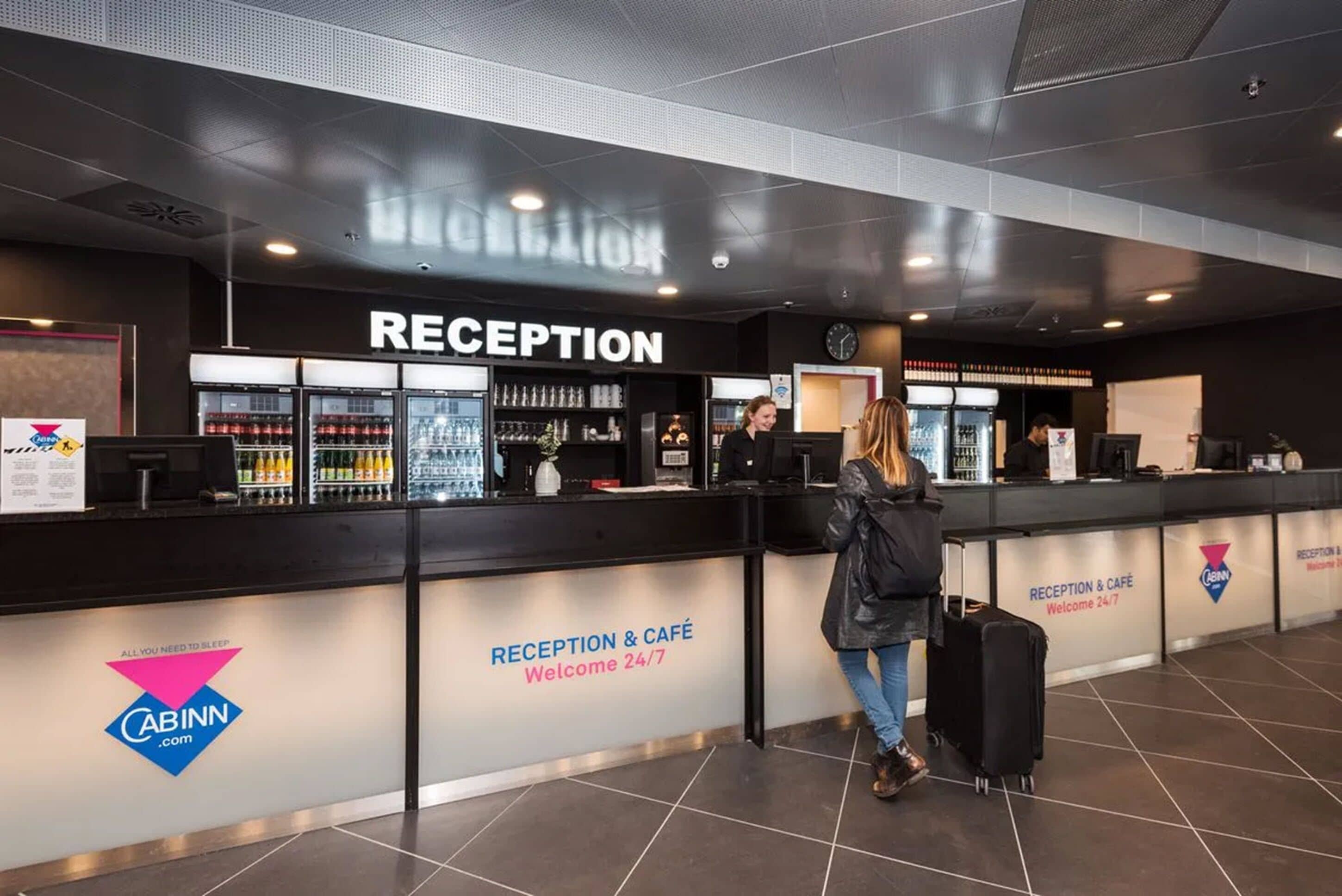 A reception area with a sign reading "RECEPTION & CAFÉ Welcome 24/7" and a logo for "CABINN.com".