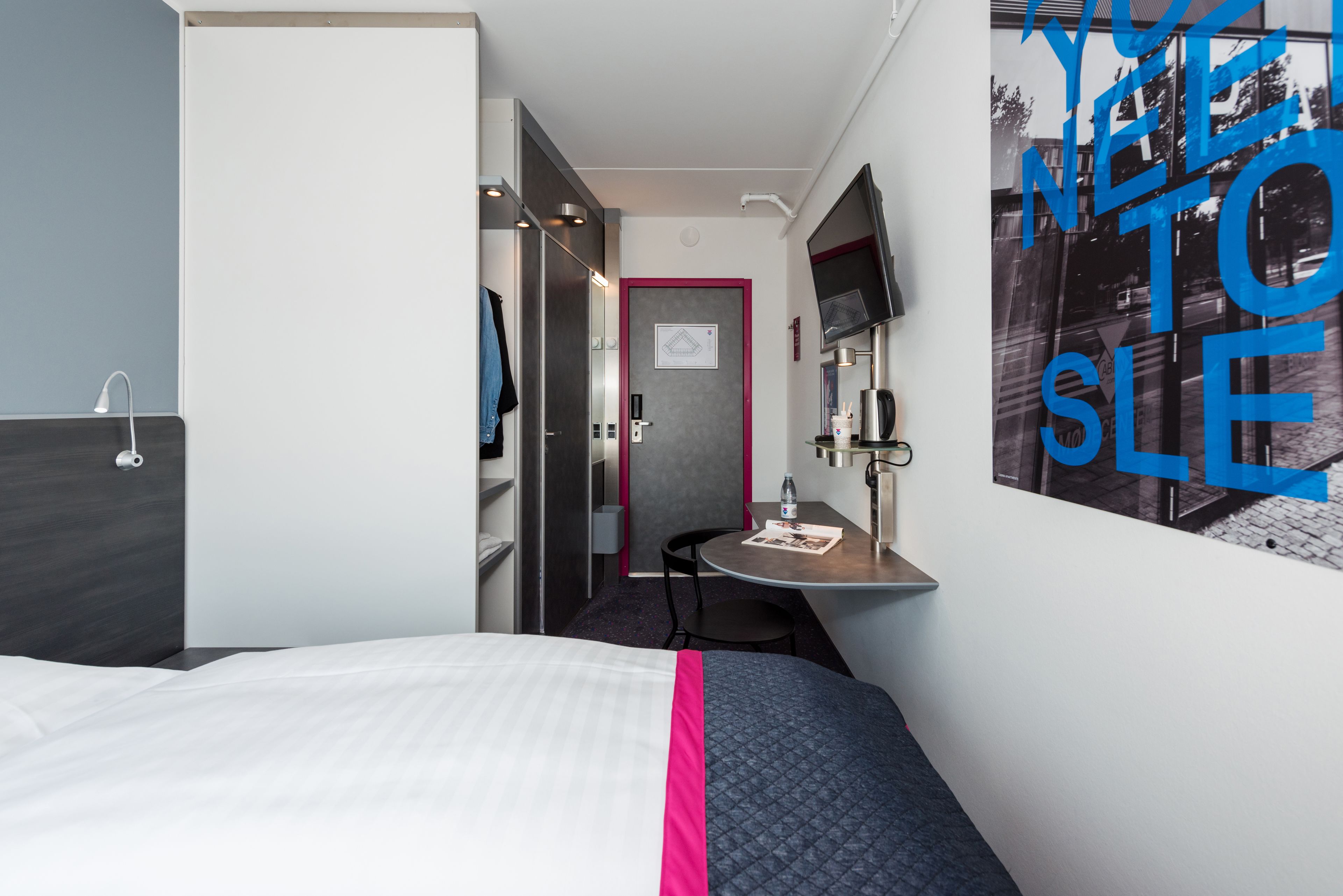 A hotel room with a bed, a desk, a chair, a mirror, and a door.
