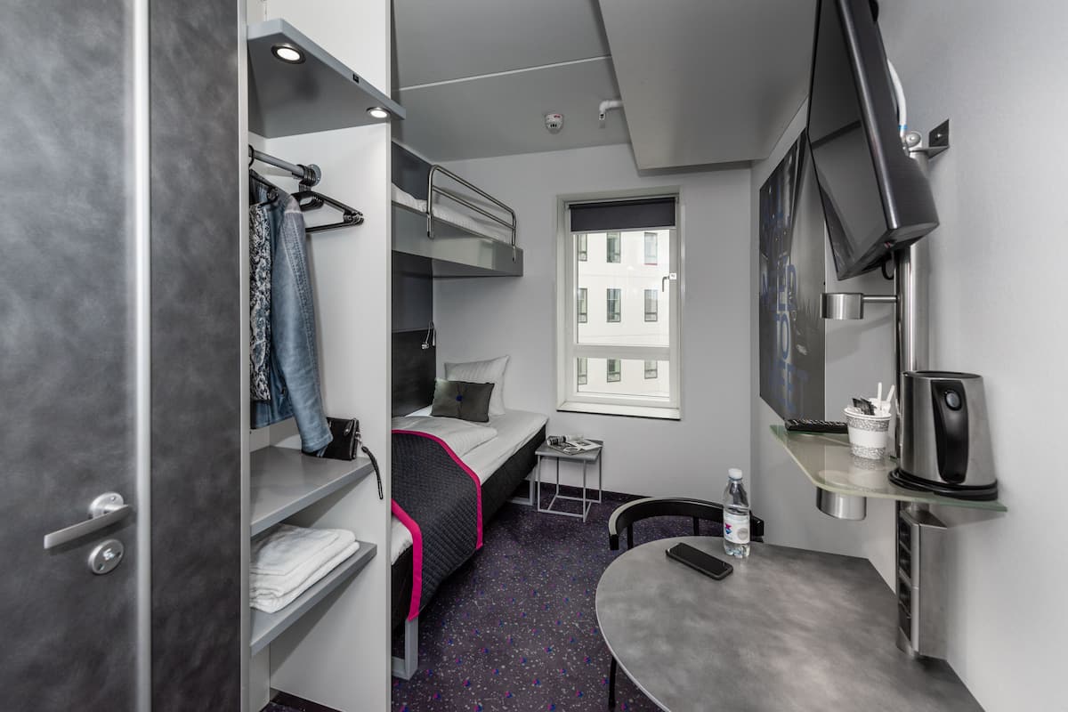 A compact hotel room with bunk beds, a small table, a chair, a TV, and a window with a city view.