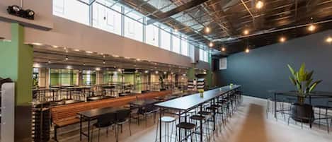 A spacious cafeteria with long tables and high ceilings, featuring industrial-style lighting and large windows.