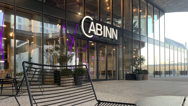 Un edificio moderno con un letrero 'CABINN', asientos exteriores con sillas de metal y grandes ventanales.