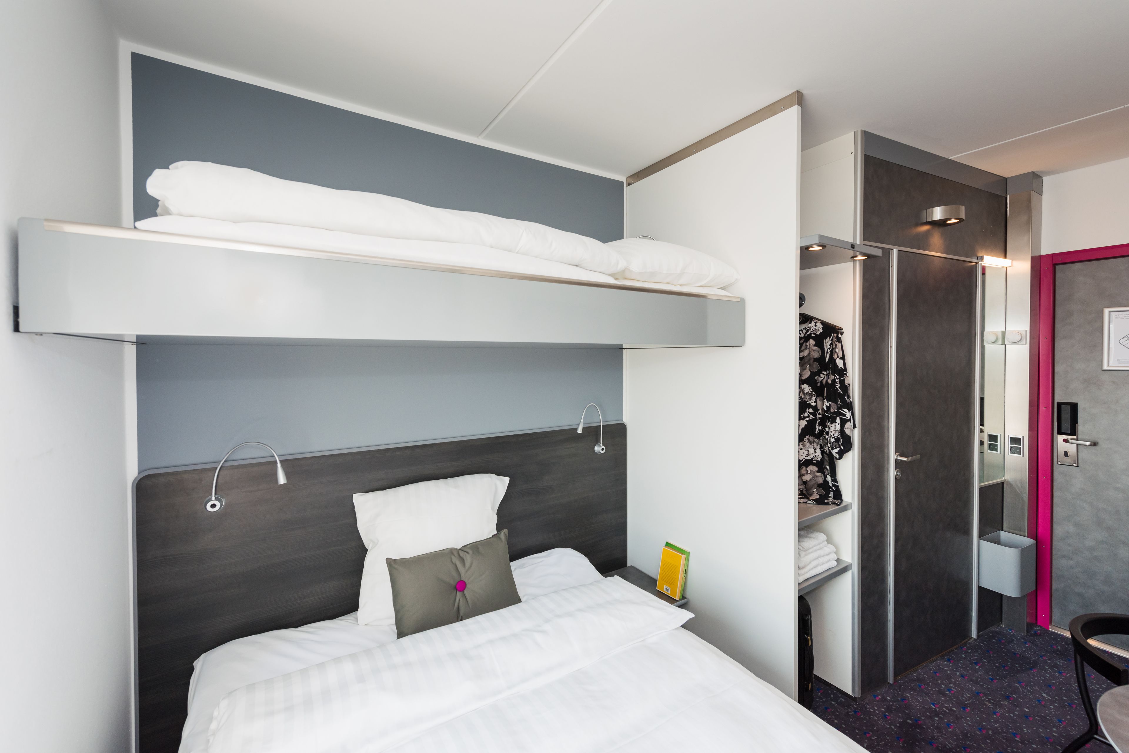 Een hotelkamer met een stapelbed, een nachtkastje met een boek, een douche en een badkamer met spiegel en handdoekrek.