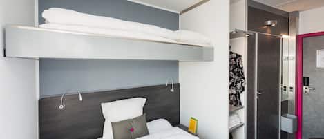 A hotel room with a bunk bed, a bedside table with a book, a shower area, and a bathroom with a mirror and towel rack.