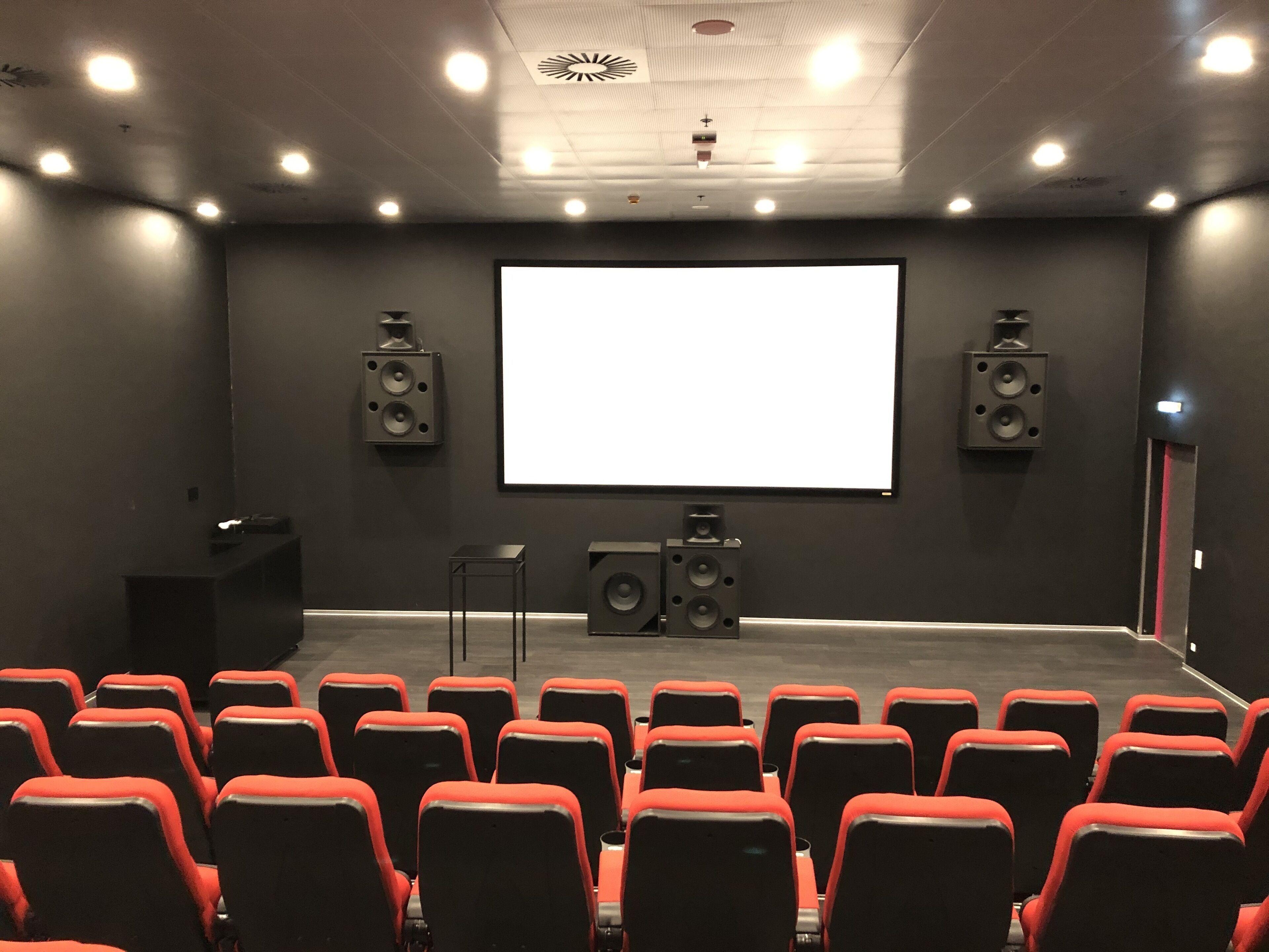 Un teatro con hileras de asientos rojos y negros, una pantalla grande y altavoces a ambos lados.