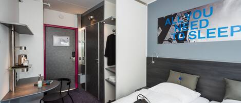 Une chambre d’hôtel moderne avec un lit, un bureau et une chaise. Il y a une affiche accrochée au mur sur laquelle on peut lire : « VOUS AVEZ BESOIN DE DORMIR ».