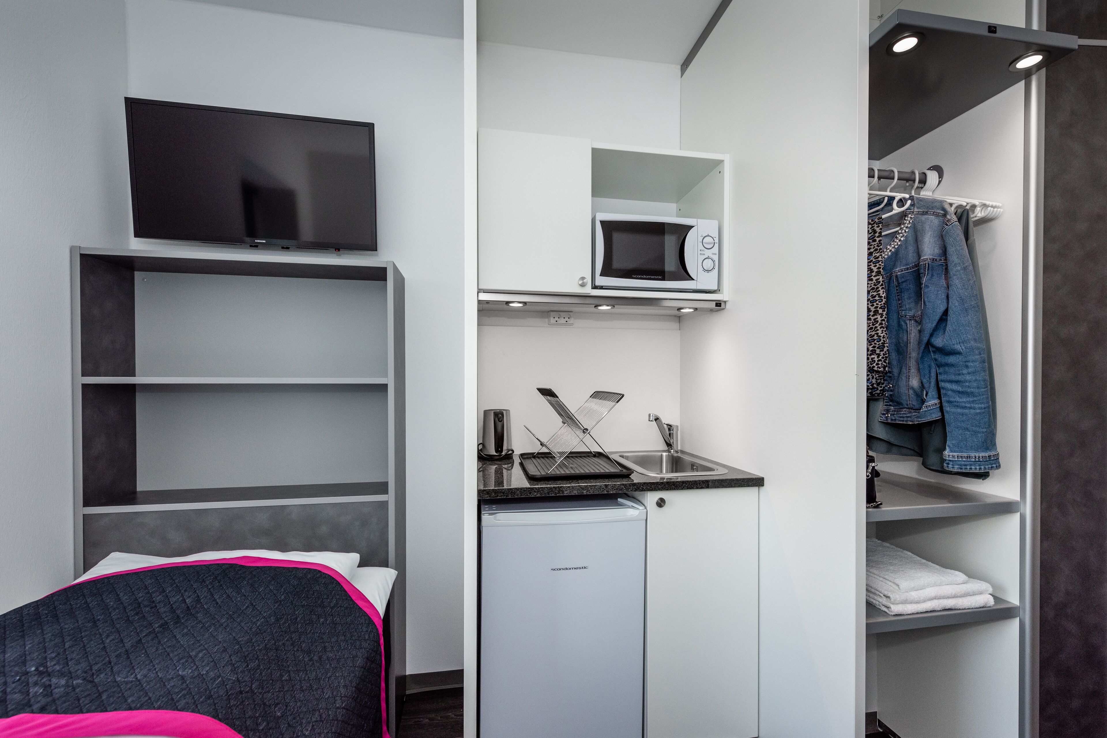 A compact room with a bed, a TV, a microwave, a small fridge, a sink, and a clothing rack.