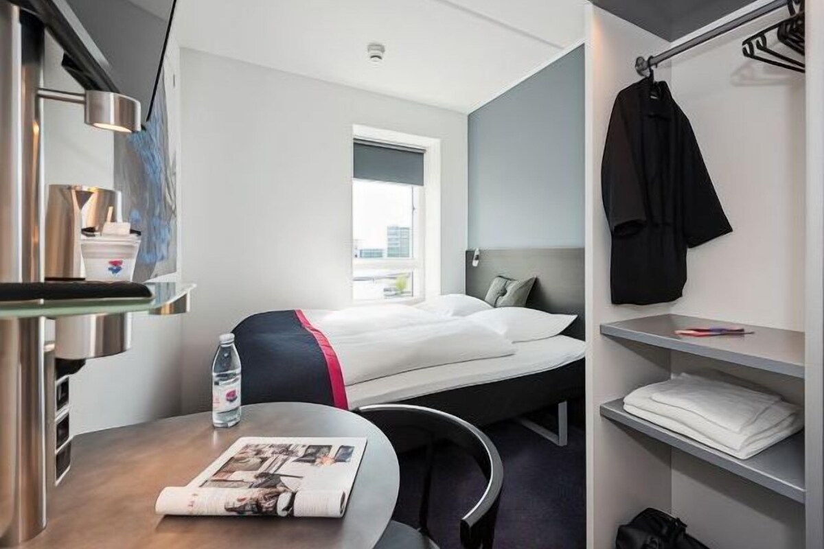 A hotel room with a bed, a desk with a newspaper, a water bottle, a coat rack, and a window.