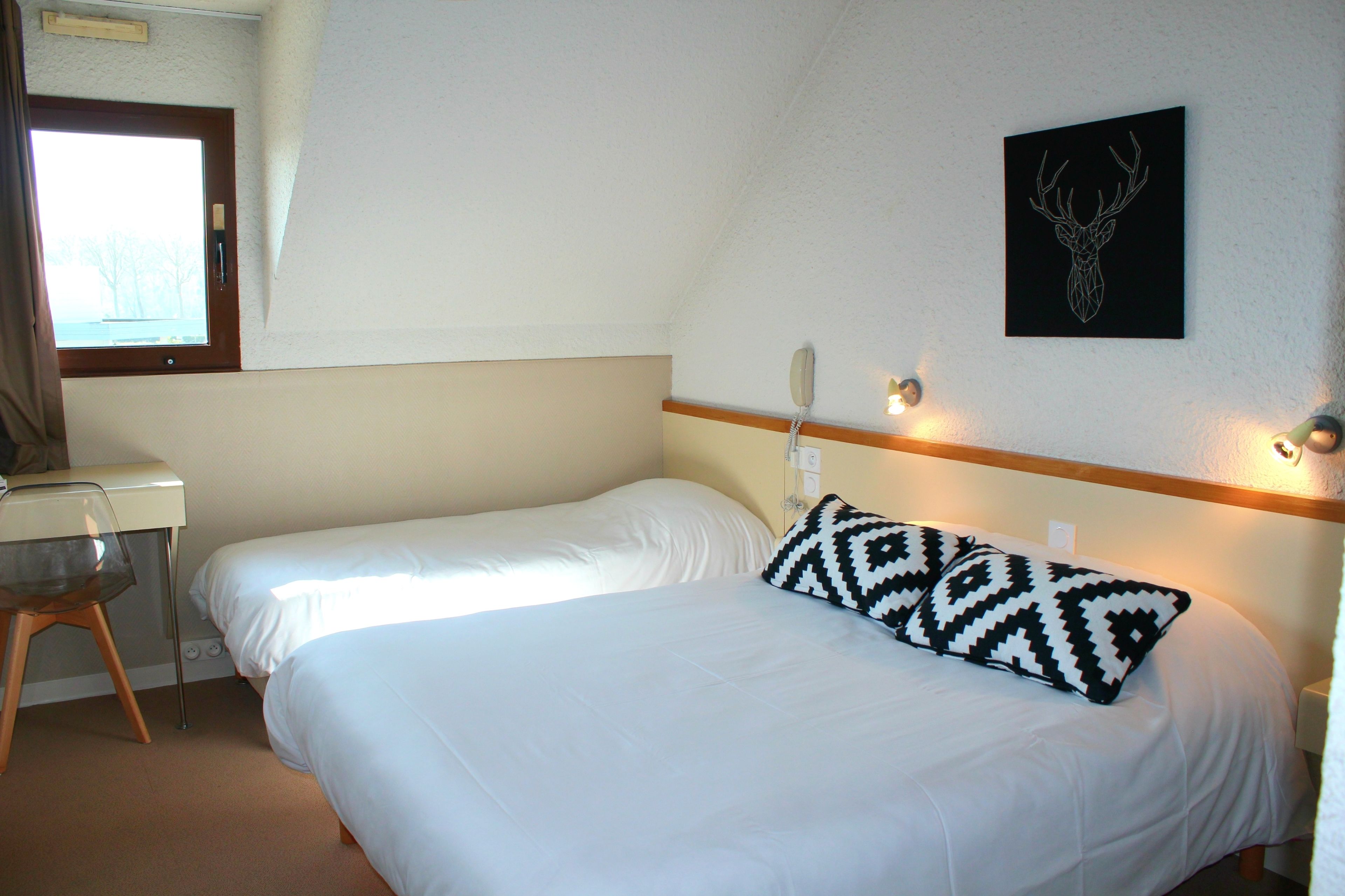 A hotel room with two single beds, a desk, a chair, a wall-mounted phone, and a deer head artwork.