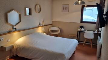 Een hotelkamer met een groot bed, een bureau met stoel, een klein rond tafeltje, een spiegel en een plafondlamp.
