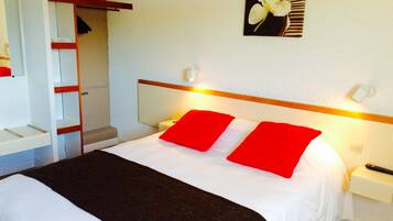 A hotel room with a bed, a nightstand, a bedside table, a lamp, a mirror, a shelf, and a door.