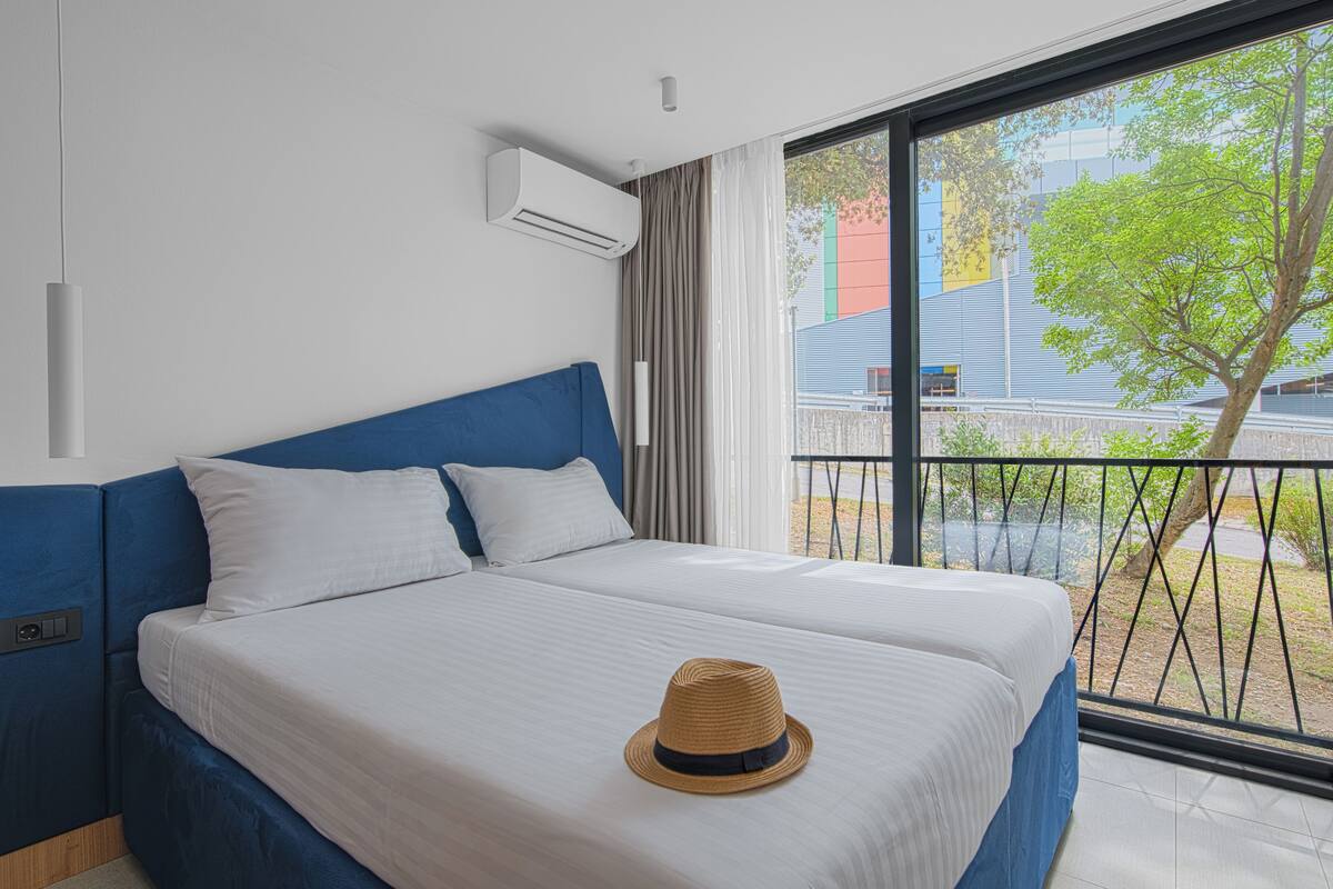 A modern hotel room with a large bed, a balcony with a view of the city, and an air conditioning unit on the wall.