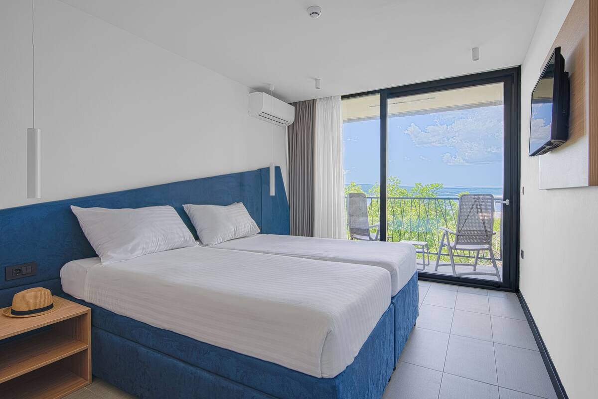 A hotel room with a large bed, a small table, a TV, and a balcony with a view of the sea.