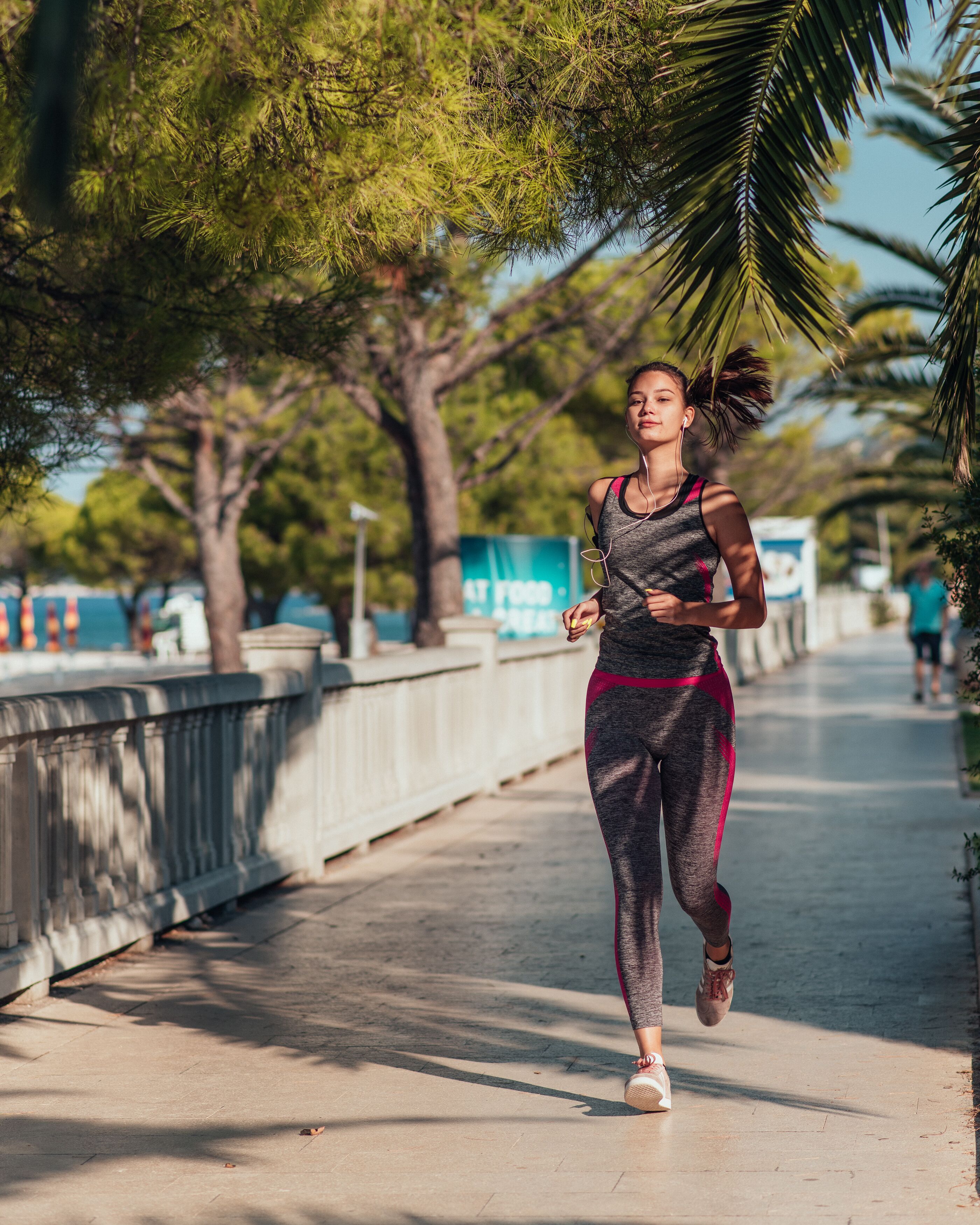 A person jogging on a paved path with trees and a railing on one side.