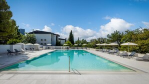 Ein rechteckiger Swimmingpool mit klarem, blauem Wasser, umgeben von weißen Liegestühlen und Sonnenschirmen, vor einer Kulisse aus Bäumen und blauem Himmel.