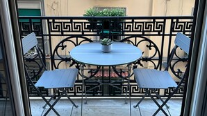 Un balcón con una mesa redonda y dos sillas, una planta en maceta sobre la mesa, y una ventana con persiana verde.