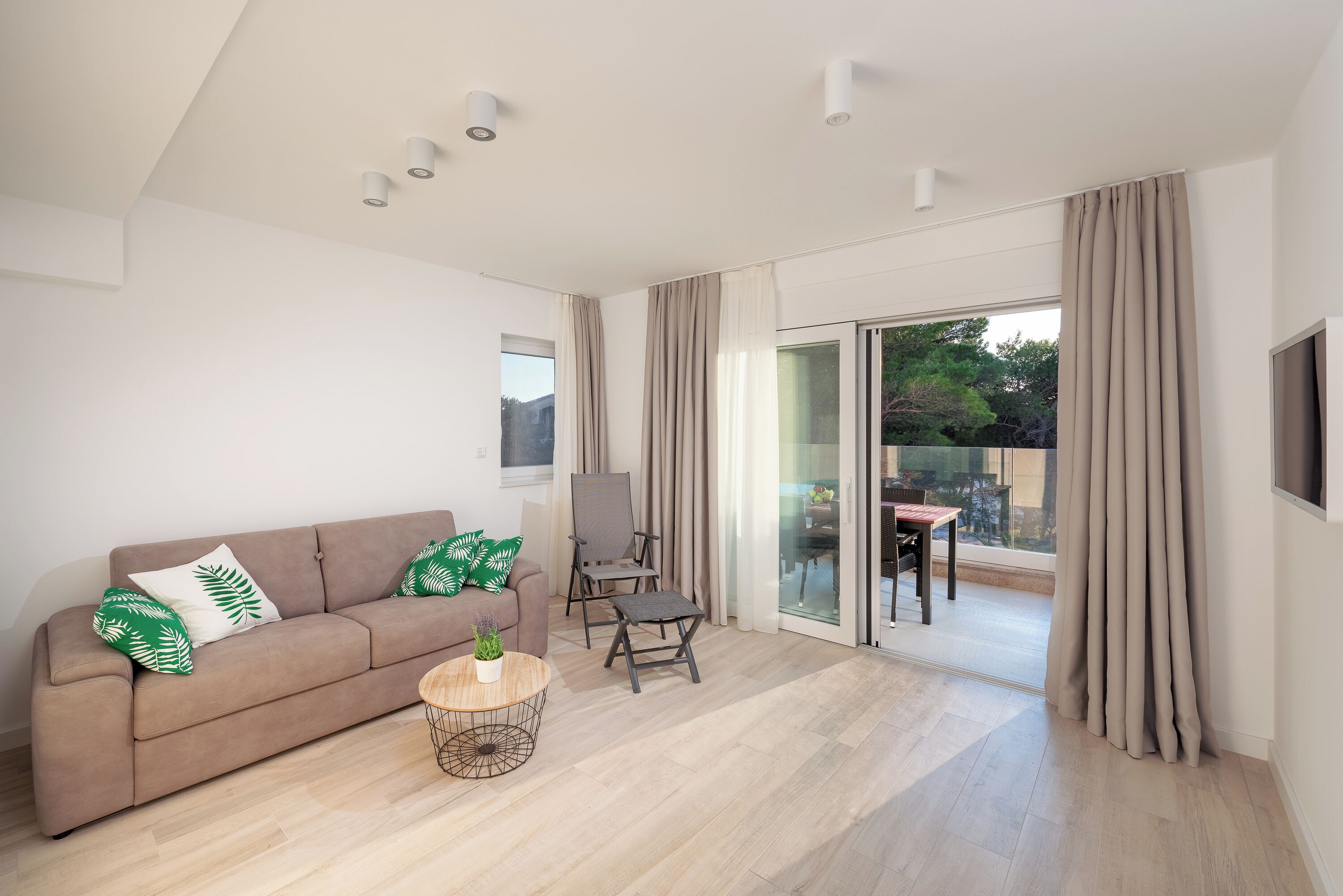 A modern living room with a sofa, armchair, and coffee table, a sliding glass door leading to a balcony, and a flat-screen TV mounted on the wall.