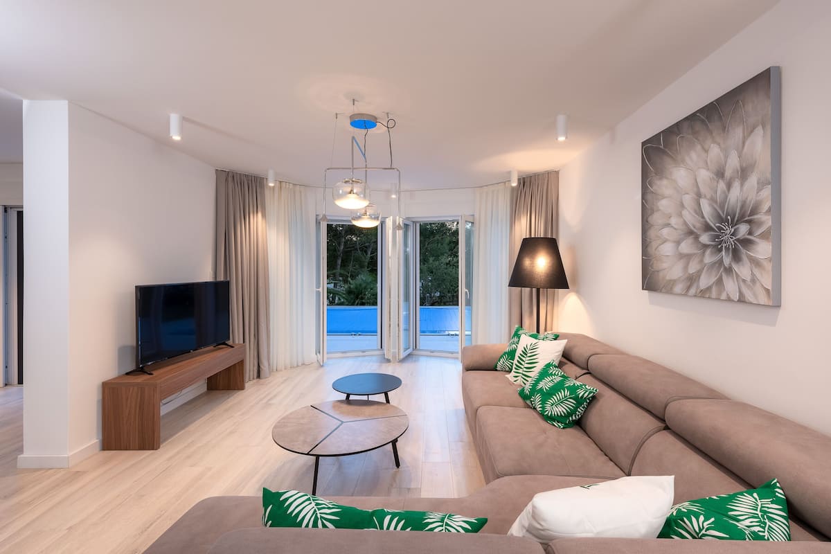 Een moderne woonkamer met een flatscreen televisie, een bank met groene en witte kussens, een ronde salontafel en een groot abstract schilderij aan de muur.