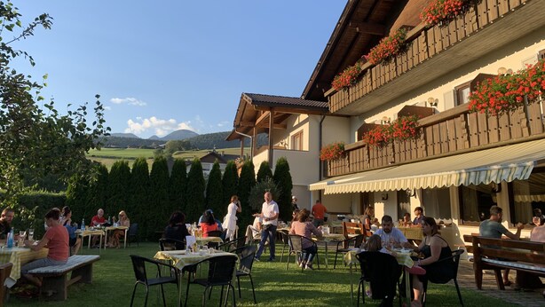 An outdoor dining area with tables and chairs, surrounded by a garden and a building with wooden balconies. - Pension Volgger (Rodengo)