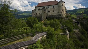 A historic castle with a round tower, surrounded by lush greenery and a winding wooden bridge. - Pension Volgger (Rodengo)